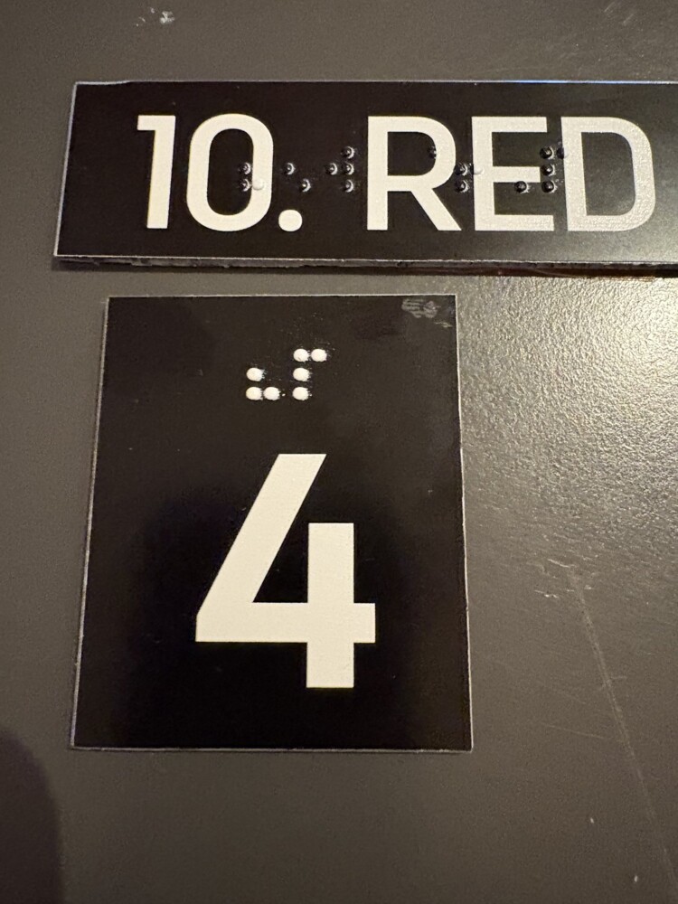 Oznaka 10. reda i sjedala broj 4 uvećanim bijelim brojkama na crnoj podlozi i Brailleovim pismom